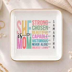Puede incluir: Un plato cuadrado de cerámica blanca con un borde dorado. El centro presenta un texto colorido que dice "SHE is MOM" junto a "STRONG", "CHOSEN", "BEAUTIFUL", "CAPABLE", "VICTORIOUS", "NEVER ALONE" y "GREATLY LOVED" con el nombre "SAMANTHA".