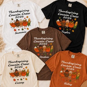 May include: Five short-sleeved t-shirts in white, black, brown, cream, and orange. Each shirt features the text "Thanksgiving Cousin Crew 2026" above a graphic of five cartoon food characters, with individual names printed below the graphic.