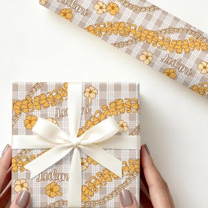 May include: A wrapped gift with a white ribbon, featuring a floral and plaid pattern in beige and yellow. The wrapping paper has the word "Leilani" printed on it. The gift is held by hands with manicured nails.