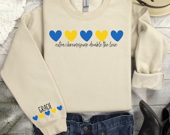 May include: A beige sweatshirt with a blue and yellow heart design and the text "extra chromosome double the love". The sweatshirt has a blue and yellow heart design on the sleeve with the name "Gracie" in black text.