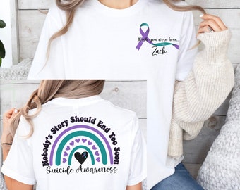 May include: White t-shirt with a purple and teal ribbon on the front that says "Wish you were here... Zach". The back of the shirt has a rainbow with a heart in the center and the text "Nobody's Story Should End Too Soon Suicide Awareness".
