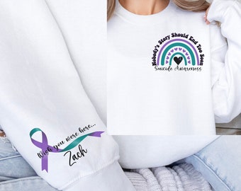 May include: White sweatshirt with a rainbow design and the text "Nobody's Story Should End Too Soon Suicide Awareness." The sleeve features a teal and purple ribbon with the text "Wish you were here... Zach."