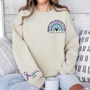 Può includere: Una felpa beige chiaro con una grafica arcobaleno viola e turchese sul davanti. L'arcobaleno ha un cuore nero al centro e il testo "Nobody's Story Should End Too Soon" e "Suicide Awareness". La manica sinistra ha un nastro viola e turchese con il testo "Wish you were here... Zach".