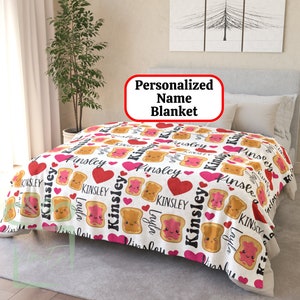 May include: Personalized blanket with a white background and a repeating pattern of cartoon toast with peanut butter and jelly, red hearts, and the name "Kinsley" in black script.