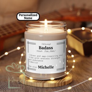 Puede incluir: Una vela de vidrio marrón con una etiqueta blanca que dice "Badass" y define la palabra como "alma valiente e inspiradora, alguien con fuerza y resistencia infinitas, guerrero". La etiqueta también dice "Ver también: Michelle".