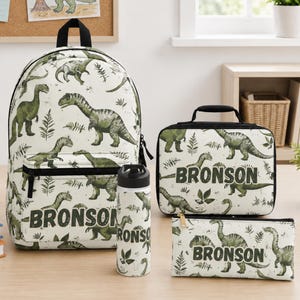 May include: A matching dinosaur-themed school set featuring a backpack, lunch bag, water bottle, and pencil case. The set is white with green dinosaur illustrations and the name "BRONSON". A drawing of a dinosaur is pinned to a corkboard.