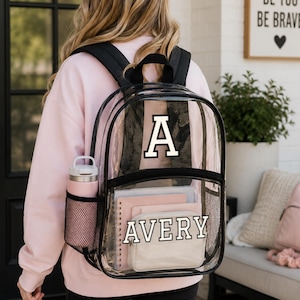 May include: Transparent backpack with black edging, displaying a white letter "A" and the name "AVERY" in white. It has a pink water bottle in the side pocket and contains pink notebooks and a small pouch. The backpack is worn by a person wearing a pink sweatshirt.
