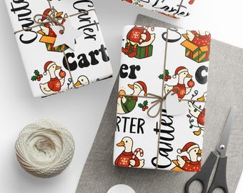 May include: White wrapping paper with a repeating pattern of cartoon ducks in Santa hats and sweaters, with the name "Carter" printed in black. The wrapped gifts are tied with twine and have white tags. A ball of twine and scissors are also visible.