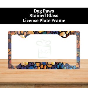 May include: License plate frame with a stained glass design featuring dog paw prints in shades of blue, yellow, and purple. The frame has the words "Dog Paws Stained Glass License Plate Frame" printed on it.