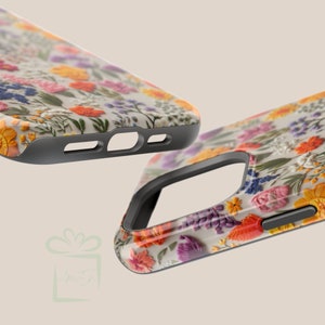 May include: A phone case with a floral design featuring embroidered flowers in shades of pink, orange, yellow, purple, and blue on a white background. The case has a black border.
