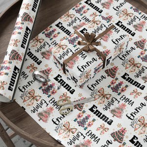 Könnte beinhalten: Weißes Geschenkpapier mit einem sich wiederholenden Muster aus Weihnachtsbäumen, Schleifen und dem Namen "Emma" in Schwarz. Ein verpacktes Geschenk mit braunem Band und Schleife liegt auf dem Papier, zusammen mit einer Rolle Papier, Klebeband und einer Schere.