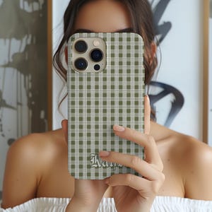 May include: A phone case with a green and white gingham pattern. The case has a camera cutout and a decorative script at the bottom. The phone case is held in front of a person's face.