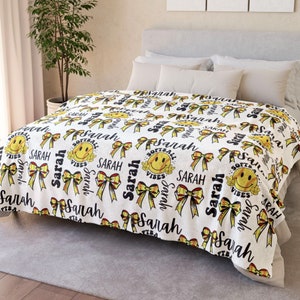 May include: White fleece blanket with a repeating pattern of yellow smiley faces wearing baseball caps, black bows, and the name "Sarah" in black text. The smiley faces have the words "Softball Vibes" written on them.