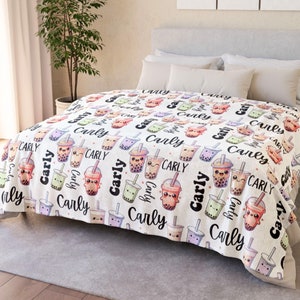 Op de afbeelding: Witte fleece deken met een herhalend patroon van cartoon bubble tea bekers met lachende gezichten. De tekst "Carly" wordt herhaald in het hele patroon.