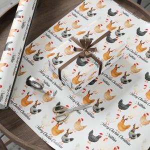 May include: White wrapping paper with a repeating pattern of chickens wearing Santa hats. A wrapped gift with a brown ribbon and bow sits on the paper. The text "Merry Cluckin Christmas Nathan" is printed on the paper.