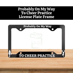May include: A black rectangular number plate surround featuring white text that reads Probably On My Way at the top and TO CHEER PRACTICE at the bottom. The frame includes white silhouettes of cheerleaders on the left and right sides.