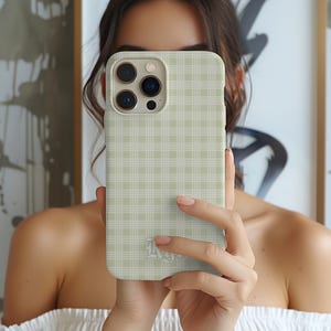May include: A light green phone case with a plaid pattern. The case has a camera cutout and a small logo at the bottom. The case is held in front of a person.
