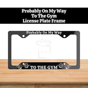 Op de afbeelding: Een zwart rechthoekig kentekenplaathouder met de witte tekst 'Probably On My Way' aan de bovenkant en 'TO THE GYM' aan de onderkant, voorzien van een afbeelding van een halter aan weerszijden van de onderste tekst in een vet lettertype.