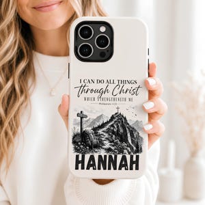 May include: A white smartphone case featuring a black and white mountain landscape illustration with a cross at the peak, a backpack, and the text I CAN DO ALL THINGS through Christ WHICH STRENGTHENETH ME Philippians 4:13 and the name HANNAH.