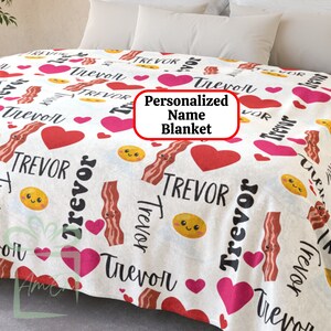 May include: White fleece blanket with a repeating pattern of red hearts, smiling bacon, and yellow eggs. The name "Trevor" is printed throughout the blanket.
