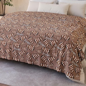 May include: A brown blanket with a repeating pattern of giraffe spots in brown and white. The word "Moani" is printed in a cursive font across the blanket. The blanket is draped over a bed with white sheets and pillows.