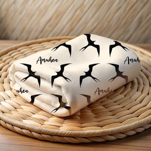 May include: A white fabric baby blanket with a black bird pattern and the word "Anuhe" repeated on the fabric. The blanket is folded and sitting on a woven basket.