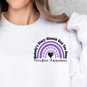 Può includere: Felpa bianca con un design a forma di arcobaleno viola e bianco. L'arcobaleno ha un cuore nero con un nastro viola al centro. Il testo "Nobody's Story Should End Too Soon" è scritto intorno all'arcobaleno. Il testo "Overdose Awareness" è scritto sotto l'arcobaleno.