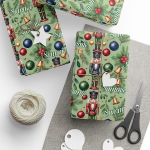 May include: Christmas-themed gift wrap featuring a repeating pattern of nutcrackers, ornaments, bells, and greenery on a light green background. The word "Sammie" is printed on the wrapping paper. The gifts are tied with twine and adorned with white heart-shaped tags.