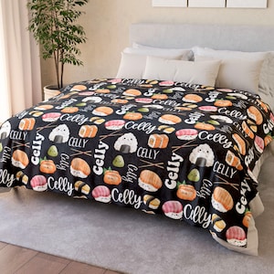 Op de afbeelding: Zwarte en witte fleece deken met een herhalend patroon van sushi-rollen, avocado, rijstballen en eetstokjes. De tekst "Celly" wordt herhaald in het hele patroon.