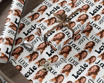 Könnte beinhalten: Weißes Geschenkpapier mit einem sich wiederholenden Muster aus einem Foto einer lächelnden Person und dem Namen "Lexie" in Schwarz. Ein verpacktes Geschenk mit braunem Band und eine Rolle passenden Papiers sind sichtbar.