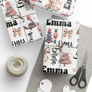 Könnte beinhalten: Weißes Geschenkpapier mit Weihnachtsmotiven, floralen Schleifen, Weihnachtsbäumen und Nussknackern. Der Name "Emma" ist fett gedruckt. Die Geschenke sind mit Bindfaden und einem herzförmigen Anhänger gebunden.