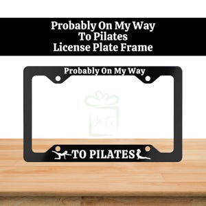 Op de afbeelding: Een zwart rechthoekig kentekenplaathouder met de witte tekst Probably On My Way aan de bovenkant en TO PILATES aan de onderkant. Het onderste gedeelte bevat twee witte silhouetten van een persoon die verschillende yoga- of pilateshoudingen aanneemt.