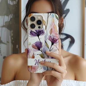 May include: A phone case featuring a watercolor-style floral pattern with purple and pink blossoms on a white background. A horizontal white band across the lower section displays the name Hailey in a purple script font. The case has a camera cutout.