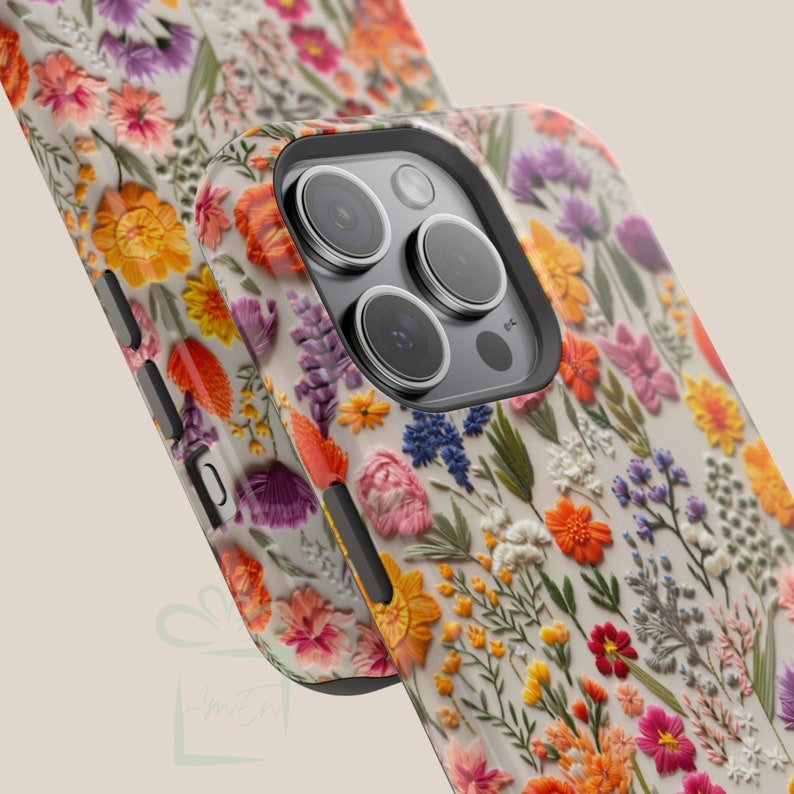 May include: A white phone case with a floral embroidery design. The case features a variety of colorful flowers and leaves, including pink, yellow, orange, purple, and green. The case has a black camera bump with three lenses.
