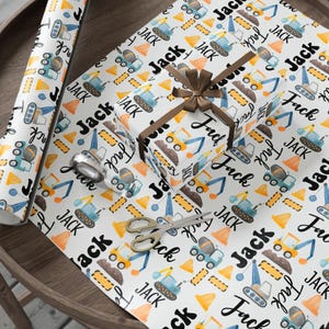 Könnte beinhalten: Weißes Geschenkpapier mit einem Konstruktionsthema. Das Papier zeigt Illustrationen von Baufahrzeugen, Leitkegeln und dem Namen "Jack" in Schwarz. Eine verpackte Geschenkbox mit braunem Band und Schere liegt auf dem Papier.