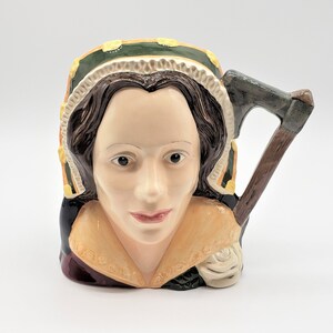 May include: A ceramic mug shaped like a woman's head, featuring a detailed face with brown hair and a decorative hat. The mug has a handle shaped like an axe. The color palette includes cream, green, yellow, and brown.
