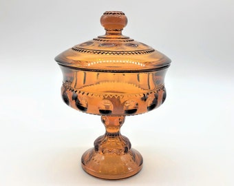 May include: Amber-colored glass candy dish with a lid. The dish has a pedestal base and a round, lidded top. The glass has a decorative pattern around the rim and on the lid. The dish is likely vintage and is suitable for serving candies or other small items.