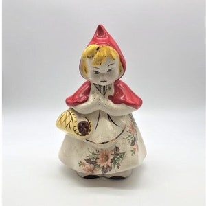 May include: A ceramic figurine of Little Red Riding Hood, featuring a red hooded cape, a white dress with floral patterns, and a yellow basket. The figurine has blonde hair and is in a kneeling position.