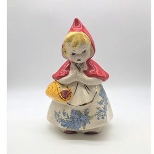 May include: A ceramic figurine of a girl in a red hooded cape, holding a yellow basket. The girl's dress is white with blue floral patterns. The figurine has a vintage aesthetic.