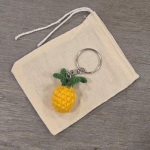 May include: A small, yellow, crocheted pineapple keyring with green leaves attached to a silver metal ring. The keyring is placed on a square, off-white, fabric drawstring pouch made of natural cotton material with two white strings at the top.