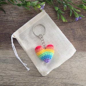 May include: A crochet heart keyring featuring horizontal stripes in red, orange, yellow, green, blue, and purple yarn. The heart is attached to a silver-toned metal split ring by a short chain. The keyring is placed on a small, off-white fabric pouch.