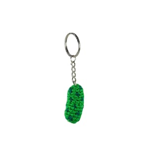 Puede incluir: Un pequeño llavero de gominola de ganchillo con una textura de hilo verde vibrante y motas de color verde oscuro. La forma cilíndrica, texturizada y acolchada está unida a una anilla de metal plateado mediante una cadena corta de eslabones.