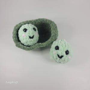May include: Two crocheted pea pods with smiling faces. One pod is inside a green pod, and the other is separate. The peas have black eyes and pink cheeks. The pods are made of a soft, textured yarn.