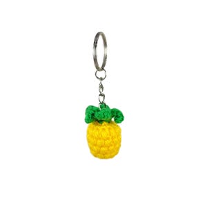 Puede incluir: Un llavero de piña de ganchillo amarillo con hojas verdes unidas a un anillo de llave plateado.