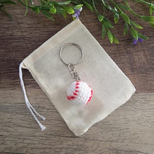 May include: A white crocheted baseball keychain with red stitching details attached to a silver metal split ring, presented on a small, plain, light beige fabric drawstring pouch with white cotton strings.