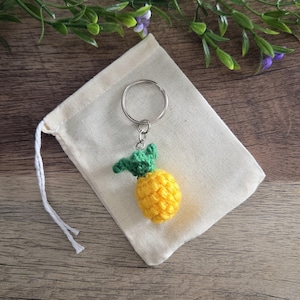 May include: A crocheted pineapple keychain featuring a bright yellow textured body and a green leafy top, attached to a silver-toned metal split ring. The small pineapple charm is crafted with yarn and sits against a plain off-white fabric pouch.