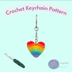 Peut inclure: Un porte-clés cœur en crochet présentant un dégradé de couleurs arc-en-ciel avec du fil rouge, orange, jaune, vert, bleu et violet. Le cœur est fixé à une chaîne et un anneau en métal argenté. Un crochet à manche vert foncé et un logo de pelote sont visibles.