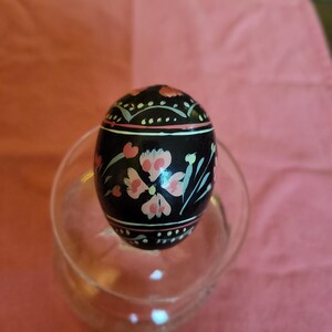 Peut inclure: Un ornement en forme d'œuf noir orné de motifs floraux peints à la main dans des tons rose pâle, vert clair et blanc. Le design comprend des bandes horizontales avec des motifs en pointillés et des fleurs centrales, posé sur une base en verre.