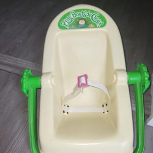 Peut inclure: Un siège porte-poupée en plastique crème avec une poignée et une base vert vif. Le siège comporte une sangle de sécurité blanche avec une boucle rose et un logo vert en haut indiquant P'tits Bouts de Choux avec une petite illustration.