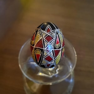 Peut inclure: Un œuf décoré présentant une base noire avec des motifs géométriques complexes en rouge, jaune et blanc. Le design se compose de formes répétitives en losange et en triangle remplies de fines hachures et de blocs de couleur unie, créant un affichage visuel détaillé, symétrique et multicolore sur la surface de l'œuf.